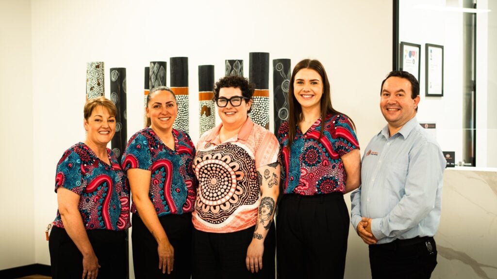 Five smiling professionals standing in a row in front of carved Aboriginal poles, with four of them wearing colourful shirts featuring Indigenous Australian art patterns.