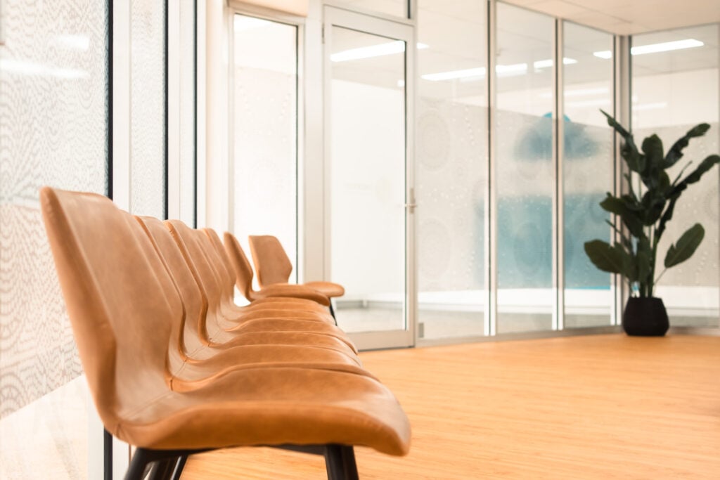 A close-up, angled view of a row of modern tan leather chairs in a bright office waiting area, with glass partitions and a potted plant in the blurred background.