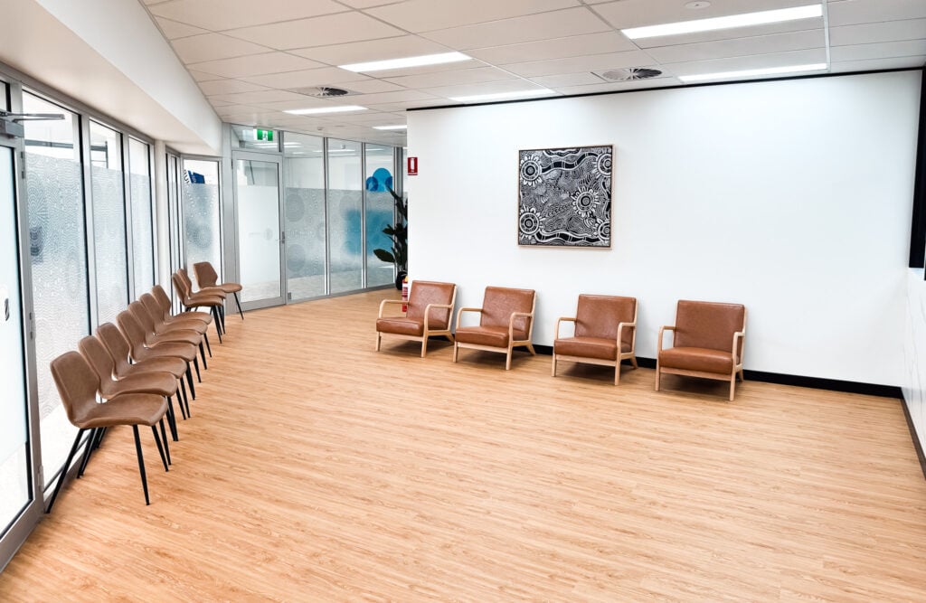 A bright, modern waiting area featuring wood-look flooring, rows of brown leather chairs, and a black-and-white Aboriginal artwork hanging on a clean white wall.