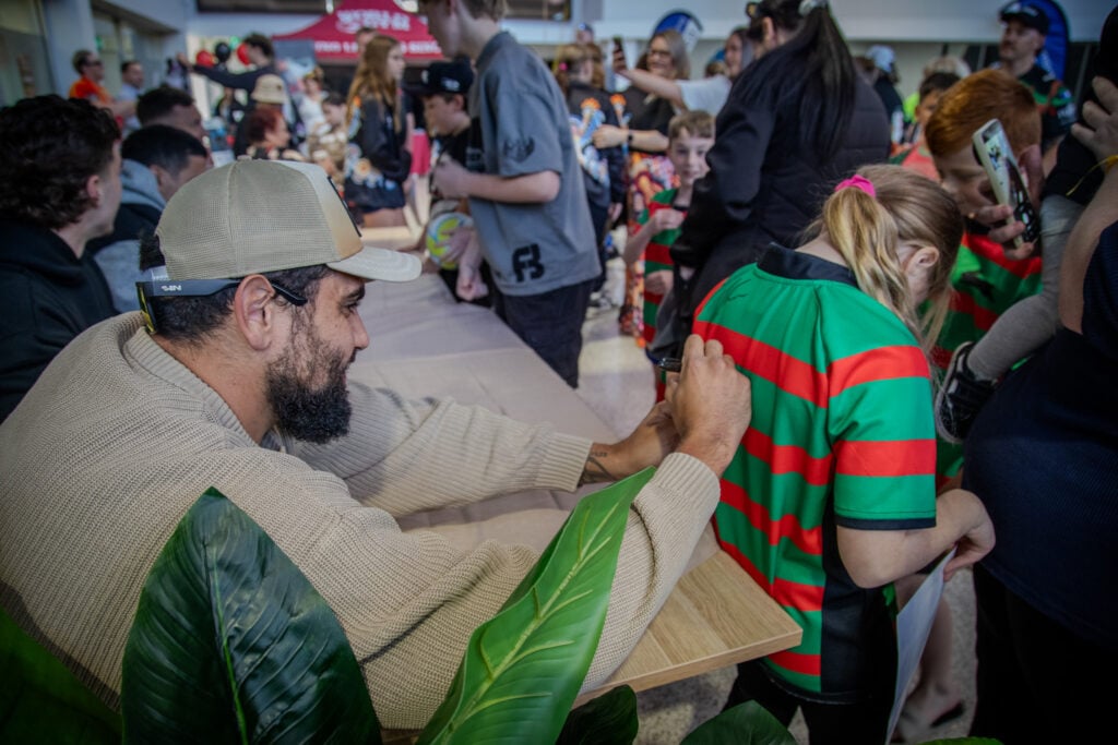 A man with a beard and a baseball cap smiling as he signs the back of a young girl's red and green striped sports jersey at a crowded community event.