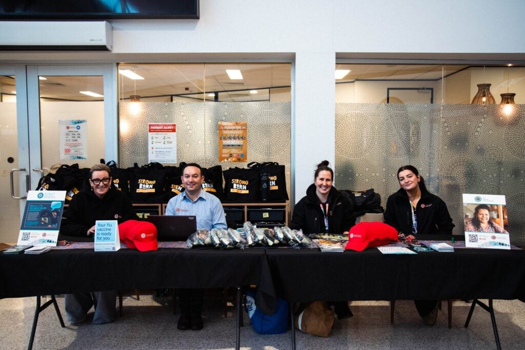 Four staff members sitting behind a promotional table at a community event, with informational posters, red hats, and black bags featuring the "Strong Born" logo on display.