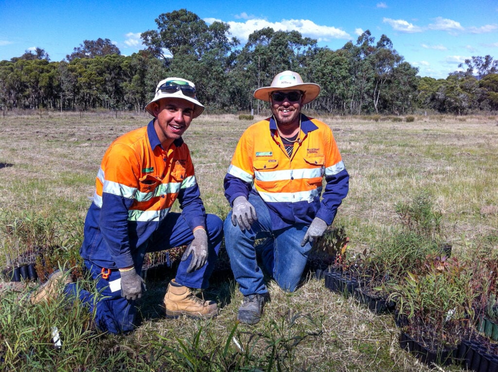 Jayden and Luke wearing high-visibility orange work shirts, wide-brimmed hats, and gloves kneeling in an open field
