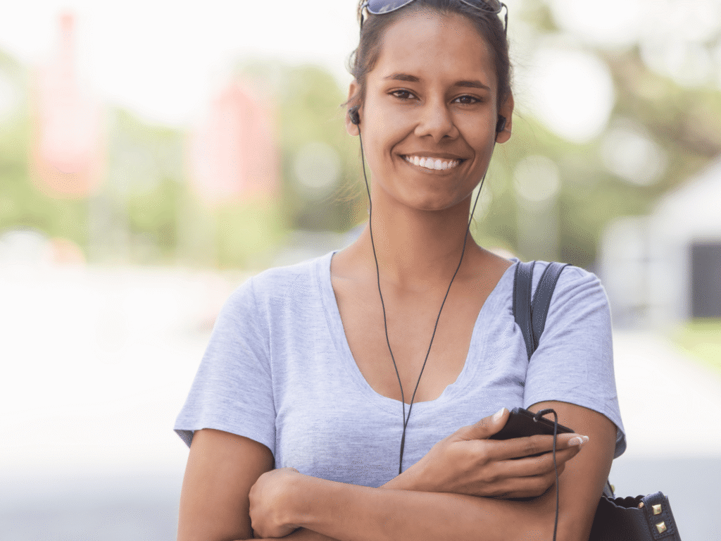 A smiling young woman wearing a grey t-shirt and wired earphones, holding a smartphone while standing outdoors